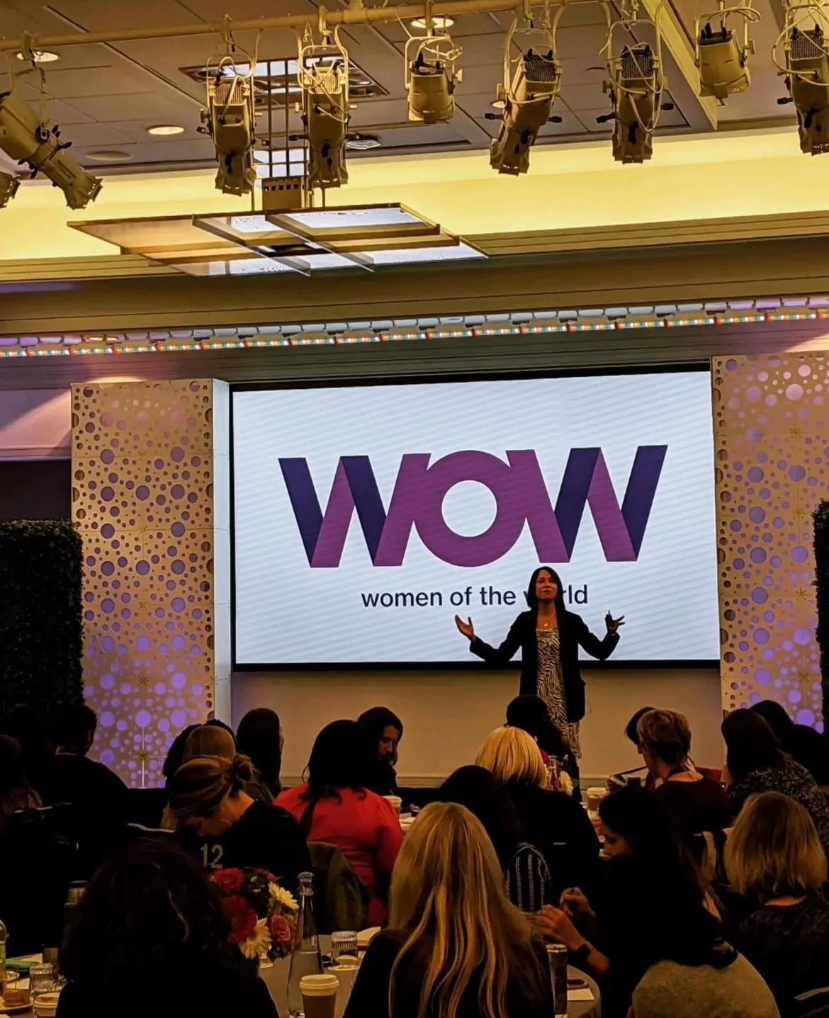 A speaker addresses a seated audience beneath a WOW women of the world screen